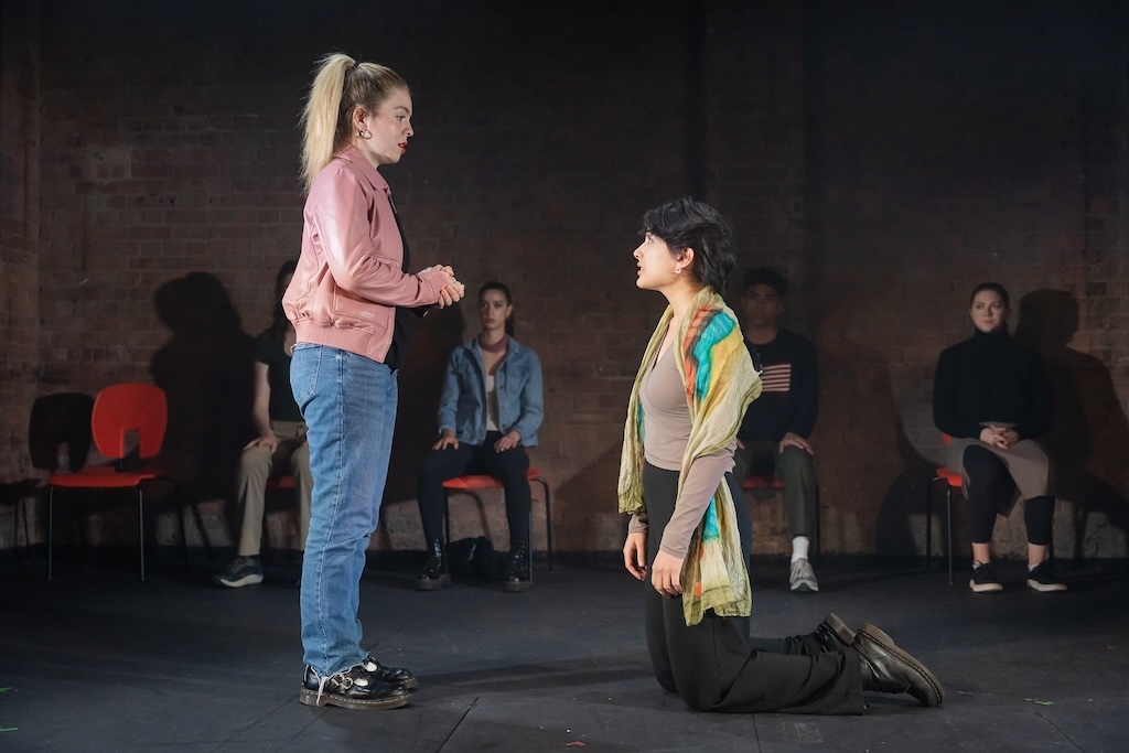 Action shot of the spring 2024 Shakespeare in Performance at RADA final presentation of Julius Caesar. Two actors in mid-performance. The actor on the right kneels in front of the actor on the left while other seated actors look on.